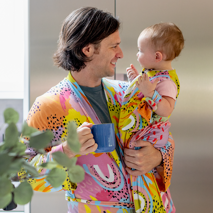Father holding baby wearing matching MiliMili x Pronoun by Jesse Tyler Ferguson Modern Rainbow Adult Robe and Wearable Blanket