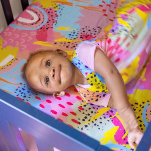 Baby wearing MiliMili x Pronoun by Jesse Tyler Ferguson Modern Rainbow Wearable Blanket on Modern Rainbow Crib Sheet, best crib sheet, bamboo crib sheet